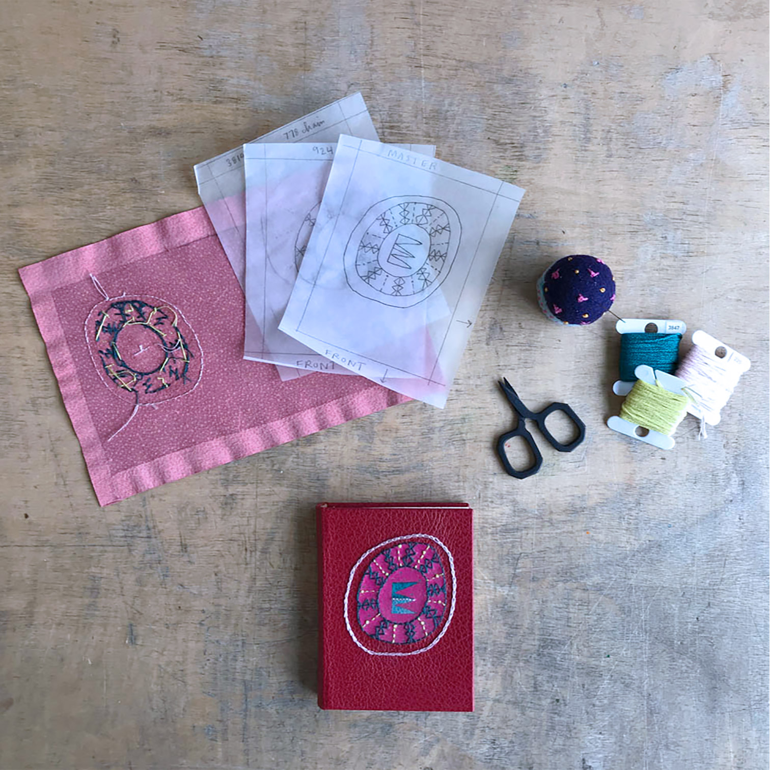 Piece of leather that has embroidery in-progress, finished and covered Embroidered Leather binding, tracing paper guides for embroidery, and bookbinding tools.